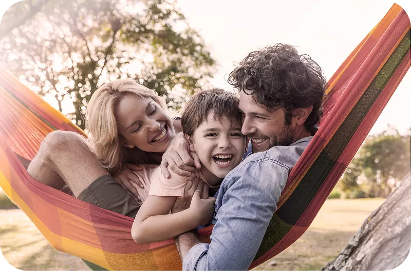 A joyful family of three—mother, father, and young son—cuddling together in a colorful hammock outdoors. All are smiling and laughing, enjoying a sunny day with trees in the background.
