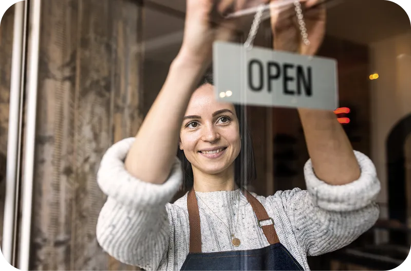 Smiling woman wearing an apron hanging an ‘Open’ sign on a glass door.